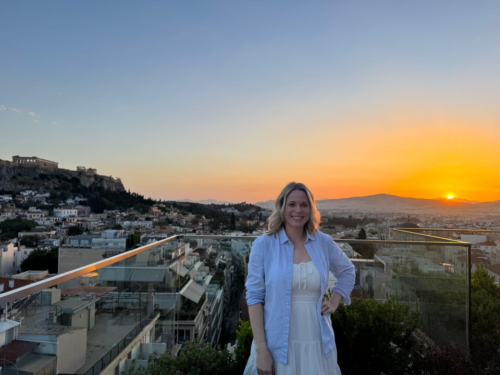 Michelle on a rooftop terrace in Athens at sunset, the Acropolis on its hill behind her